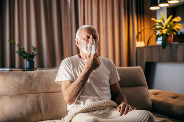 An ill senior man is sitting at home and using nebulizer.