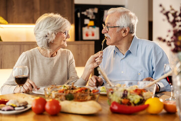 A senior woman is feeding her husband at lunch table at home.