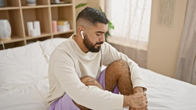 A thoughtful man with a beard sitting on a bed indoors, wearing earbuds and casual clothes.