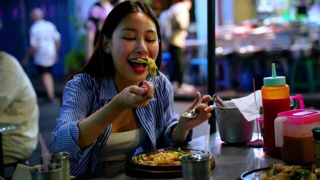 Young adult Asian woman enjoys eating a local Thai food in the street food stall shop at night in Bangkok, Thailand. Solo backpacker eating street food on table in the street.