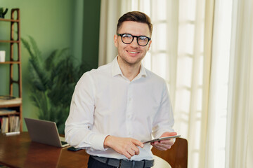Cheerful businessman in a white shirt using a tablet in a sunny room with a laptop on the desk.