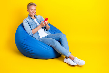 Full size photo of pretty young girl sit bean bag hold device influencer dressed stylish denim outfit isolated on yellow color background