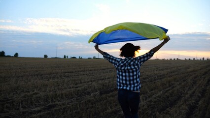 Ukrainian woman running with raised flag Ukraine above her head on wheat field at sunset. Lady jogging with national blue-yellow banner on barley meadow at sunrise. Victory against russian aggression. - 757920311