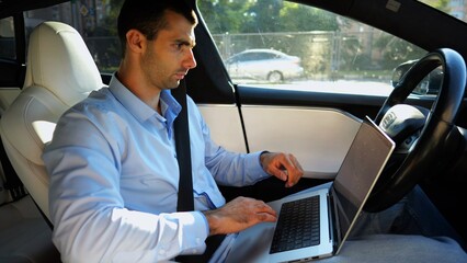 Handsome entrepreneur working on notebook while riding an autonomous self driving electric car at urban road. Male businessman typing text on laptop during riding on electrical vehicle with autopilot - 757920777