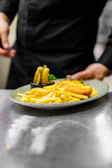 chef in black attire holding a plate with a grilled cheese chicken, fries, and garnish in a kitchen. 