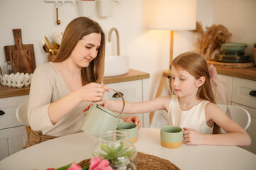 Mother pours cacao for child. Happy mother with daughter drink tea or cocoa sitting in kitchen and having fun. Family communication. Mom and children spend time together. Happy Mother's Day.