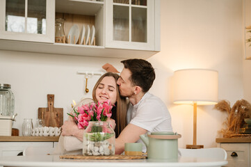 Obraz na płótnie Canvas Male kiss and hug female, spending time together. Man congratulate woman and give bouquet of flowers on Women's day. Loving couple having conversation and drinking coffee in morning in kitchen.