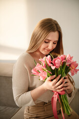 Beautiful woman is smelling a bunch of spring flowers and sitting on sofa in living room at home. Portrait young girl in dress holding in hands a big bouquet of pink tulips. International Women's Day.