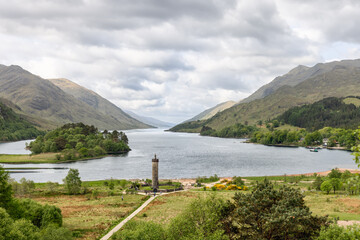 The iconic Glenfinnan Monument stands tall at the water's edge, surrounded by the lush landscape and majestic mountains of the Scottish Highlands under a moody sky