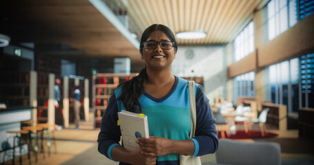 Portrait of a Cheerful Indian Student Standing in a Traditional Public Library. Young South Asian Female Looking at Camera and Smiling. Scholar Holding Academic STEM and Math Textbook