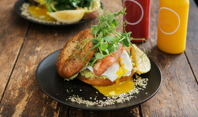 Tasty sandwich with avocado, salad and poached egg on a plate with jars of juice, close up, selective focus. Brunch on a rustic coffee shop table.