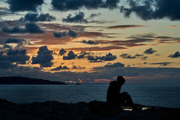 unrecognizable girl in silhouette sitting at the edge of steep and rugged cliffs of the gulf of biscay bay near St. Jean de Luz and using her smart phone with dramatic sunset and cloudscape.