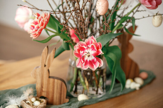 Stylish easter decoration on wooden table close up in room. Beautiful tulips bouquet with willow branches, easter eggs and wooden bunny figurines decor. Happy Easter!