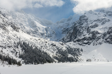 Frozne Morskie Oko Lake covered with snow in the sunny day with people on its surface. Partly cloud covered High Tatra Mountains peaks like Rysy in the background.