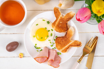 Easter breakfast or brunch with fried eggs, bacon slices and toasted bread shaped in form of bunny rabbits face, with spring flowers and Easter chocolate eggs on white wooden table copy space