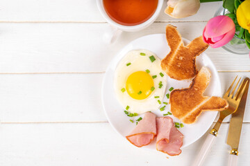 Easter breakfast or brunch with fried eggs, bacon slices and toasted bread shaped in form of bunny rabbits face, with spring flowers and Easter chocolate eggs on white wooden table copy space