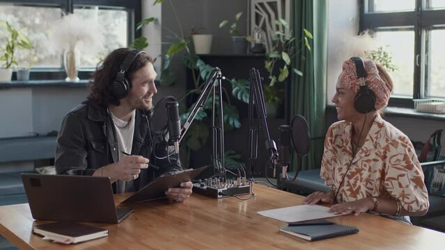 Medium footage of diverse co-hosts having warm communication while recording podcast at table with studio equipment
