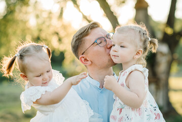 A father hugs two daughters. Daddy kisses the kids and spends time together. Dad embraces the child's girls in a park at sunset. Portrait of a daughter hugging father in a field on summer day. Closeup