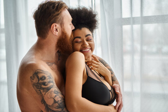 bearded and tattooed man without shirt kissing his happy african american girlfriend near curtain