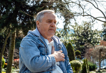 Worried elderly man experiencing chest pain while clutching his heart in park setting.