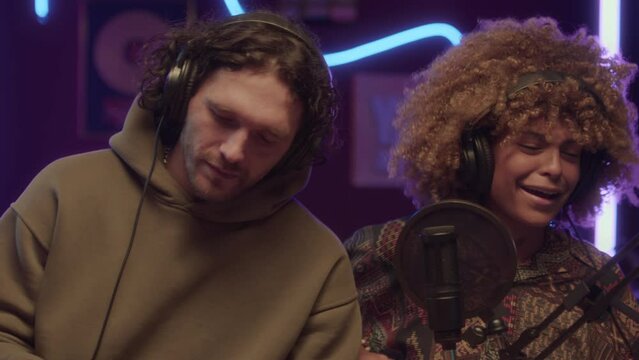 UGC chest up pan footage of smiling diverse friends recording song and moving head to music at home studio with neon string lights at night