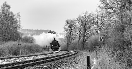 Historic passenger train with steam locomotive in a curve of the main railway line between Hagen and Arnsberg in Sauerland, Germany. Panoramic view of in winter scenery with snow and bare trees.