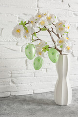 Easter eggs hanging on a decorative cherry branch, white brick wall. Side view, selective focus. Vase with cherry branch and Easter eggs. - 757977528