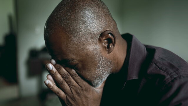 One depressed senior man covers face in shame and regret in moody gloomy bedroom. African American person struggles with poverty and mental illness, close-up face feeling hopeless