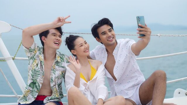 Asian man and woman friends enjoy outdoor lifestyle travel ocean on sailing luxury private catamaran boat yacht and using mobile phone taking selfie together on summer tropical holiday vacation.