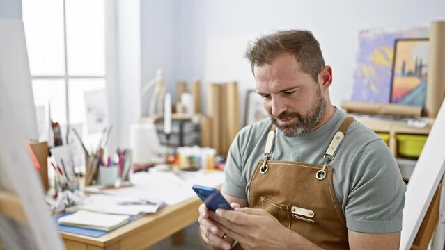Handsome middle-aged man in an apron using a smartphone in a bright art studio.