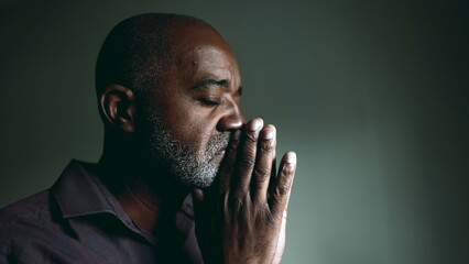 One religious black senior man in contemplative PRAYER at home in dimly lit room with eyes closed...