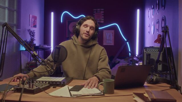 UGC of Caucasian bearded man looking at camera and talking into microphone while recording video content for vlog at home studio with neon string lights at night
