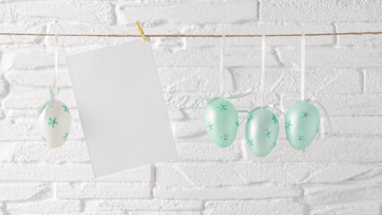 Blue Easter eggs and blank card mockup hanging on a rope, white brick background. Side view, selective focus. Banner - 757984773