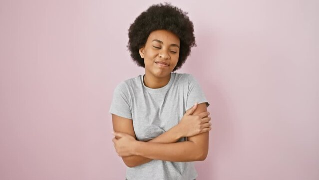 Joyful african american woman, hugging herself with love and care, confidently standing isolated on pink background, wearing a t-shirt and basking in self-love. her happy smile radiates positivity.