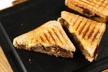 Beef toast sandwich. Close up food photo with a sandwich made of grilled bread, beef, cheese, pickles and basil on a black plate with rock and wood. Fast food photography.