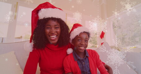 Image of snow falling over smiling mother and daugher with santa hats waving hands