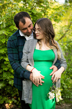 Front image of a lovely couple expecting a baby spend time outdoors on a sunny day, being hugged. Fatherhood concept.