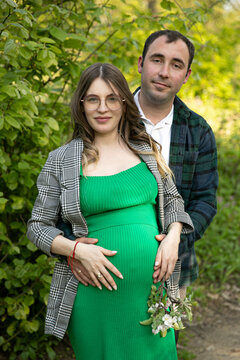 Front image of a young couple expecting a baby spend time outdoors on a sunny day, being hugged. Fatherhood concept.
