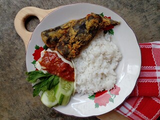 Lele krispi or crispy catfish. The catfish is seasoned and then fried in a flour coating. Served with rice, chili sauce and fresh vegetables. Top view.