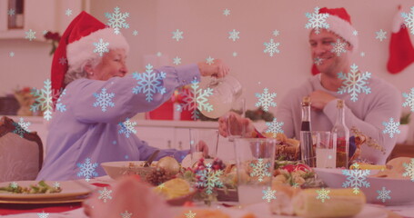 Image of snow falling over smiling caucasian family with santa hats having dinner