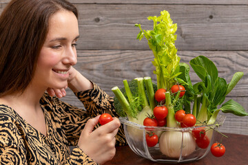 A bowl with fresh vegetables: tomatoes, celery stalks, fennel bulbs, broccoli, bok choi. A young woman holding a small tomato. Healthy vegan lifestyle concept. - obrazy, fototapety, plakaty