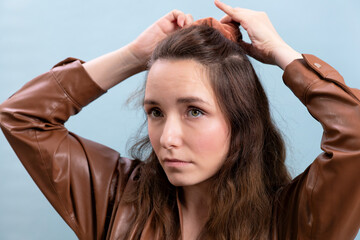 Naklejka premium Portrait of the female person in a brown eco-leather dress making hairstyle herself. A woman is preparing to go to a meeting.