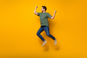 Fototapeta na wymiar Full size photo of handsome young guy raise fists winning jumping wear trendy khaki outfit isolated on yellow color background