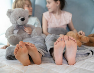 Two barefoot children, brother and sister, sitting on the bed, holding soft toys in their hands, are having fun communicating. children 7-8 years old at home on a big bed, morning rest, family joy