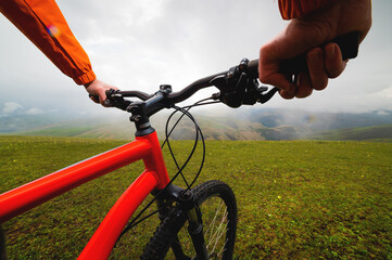 Naklejka premium Hands hold the steering wheel of a bicycle while standing on a green meadow. A look through the eyes of a biker
