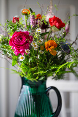 Bunter Blumenstrauss in Glasvase - Strauss aus Frühlingsbluemen - Ranunkel, Kamille, Strandflieder, Zweige, Spirea, Veronika, Distel und  in einer türkisfarbenen Glasvase vor einem hellgrauen Schrank.