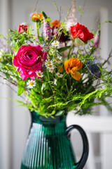 Bunter Blumenstrauss in Glasvase - Strauss aus Frühlingsbluemen - Ranunkel, Kamille, Strandflieder, Zweige, Spirea, Veronika, Distel und  in einer türkisfarbenen Glasvase vor einem hellgrauen Schrank.