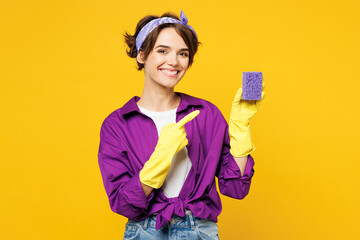 Young smiling happy woman she wear purple shirt rubber gloves do housework tidy up hold in hand point index finger on sponge isolated on plain yellow background studio portrait. Housekeeping concept. - obrazy, fototapety, plakaty