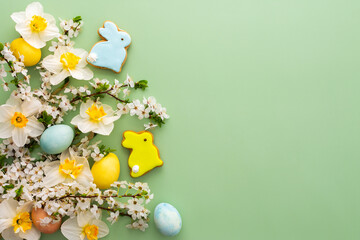 Festive background with spring flowers and naturally colored eggs and Easter bunnies, white daffodils and cherry blossom branches on a green pastel background