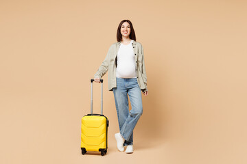 Traveler pregnant woman wears grey casual clothes hold suitcase bag ticket walk go isolated on plain beige background. Tourist travel abroad in free time rest getaway. Air flight trip journey concept. - obrazy, fototapety, plakaty